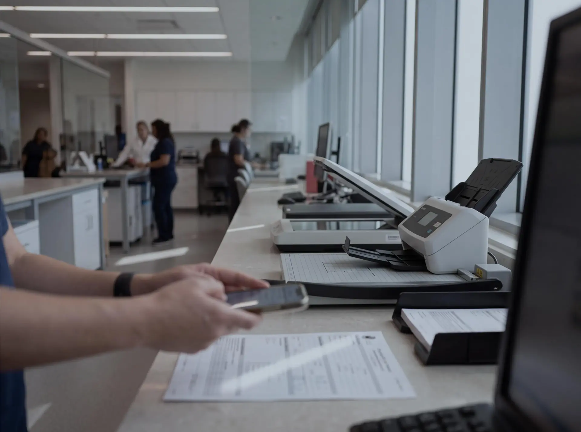 A lab technician photographing a requisition at a front-desk counter lined with document scanners